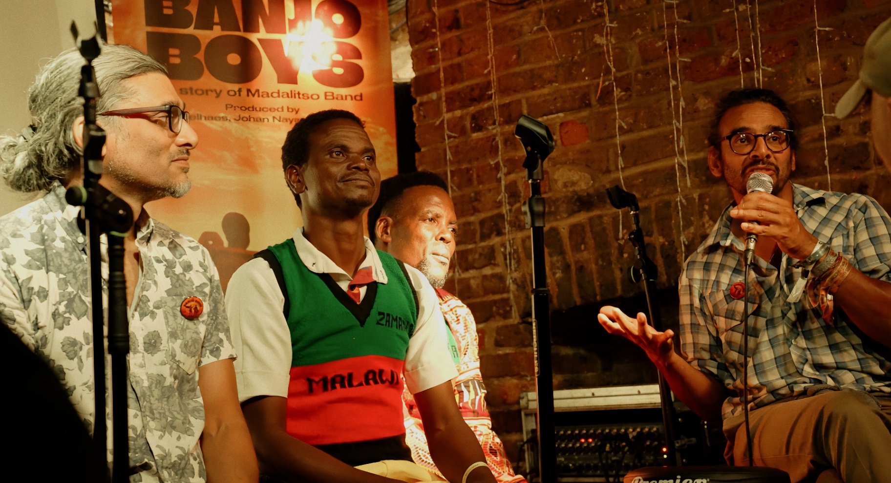 Johan Nayar, Yobu and Yosefe seated beside the Banjo Boys poster while Neil Nayar speaks into the mic at The Madalitso Experience