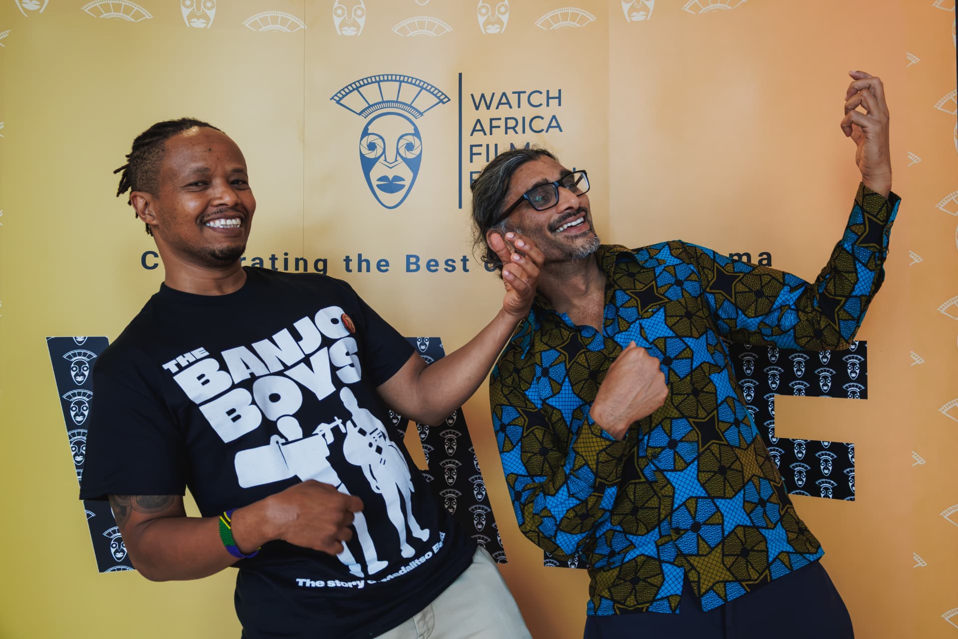 Fadhili Maghiya and Johan Nayar playing air banjos in front of the Watch Africa Film Festival backdrop at Chapter Arts Centre, Cardiff
