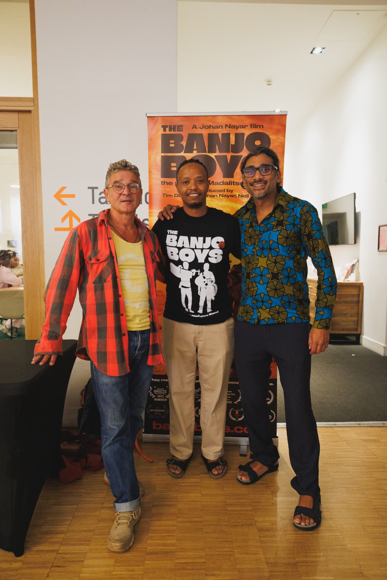 Tim Tyson Short, Fadhili Maghiya and Johan Nayar beside The Banjo Boys roll-up banner at the Watch Africa screening at Chapter Arts Centre, Cardiff